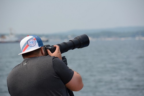 A man in a backwards baseball cap faces away from the camera. He holds a large telephoto zoom lens and is poised to take a picture.